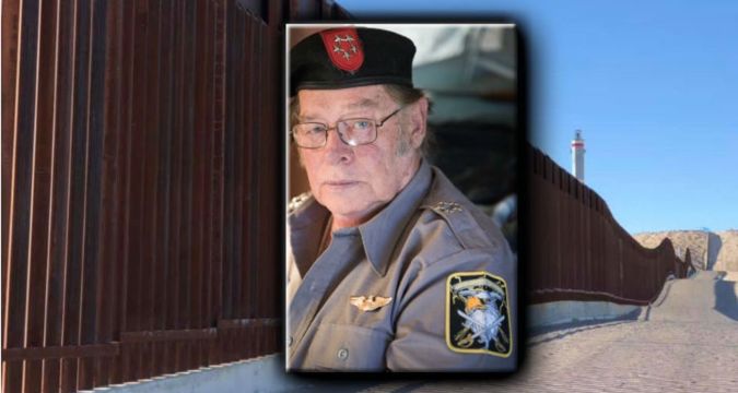 A portrait of Larry Mitchell Hopkins in a military-style uniform and beret, overlaid on a background of the US-Mexico border wall.