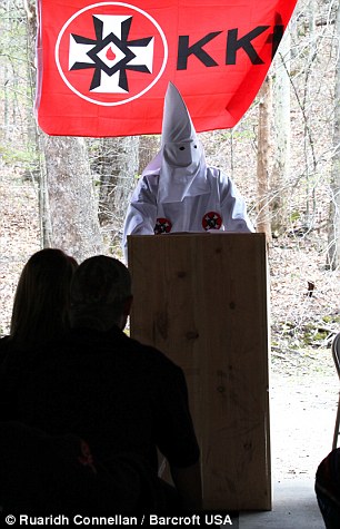 A person wearing a white Ku Klux Klan robe and pointed hood stands behind a wooden podium in a wooded outdoor setting. A red flag with the KKK insignia hangs behind them. Two people are visible from the back in the foreground.