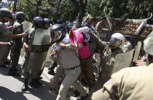 Cambodian security forces in tan uniforms and helmets forcibly detaining a protester wearing a pink shirt and yellow polka-dot pants during a street demonstration.