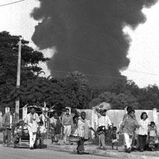 A black and white photograph showing a large plume of dark smoke rising over a line of trees and buildings, while a group of people, including children, walk along a road in the foreground, appearing to flee the scene.