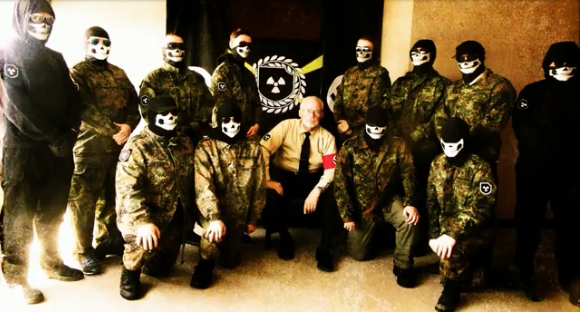 A group of individuals in camouflage uniforms and skull masks posing with a seated man wearing a tan shirt, black tie, and a Nazi swastika armband. A flag with a radiation symbol is visible in the background.
