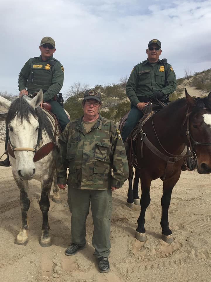 A photograph showing three men in a desert setting. Two men in green Border Patrol uniforms are mounted on horses. A third man, wearing a camouflage jacket and a cap, stands between them on the ground.