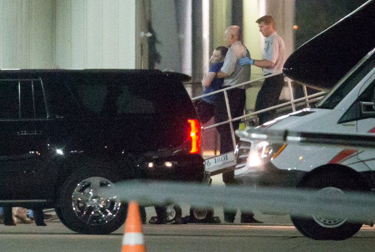 A nighttime photograph showing Otto Warmbier being carried down a ramp from a medical evacuation aircraft by two medical personnel. A black SUV and a white medical van are parked nearby on the tarmac.