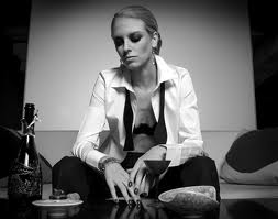 A black and white photograph of a woman sitting on a couch, wearing an open white shirt, black bra, and an untied bowtie, looking down at a table with a bottle and glass.