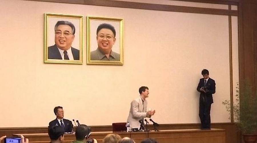 Otto Warmbier speaking at a press conference in North Korea, with portraits of Kim Il-sung and Kim Jong-il in the background.