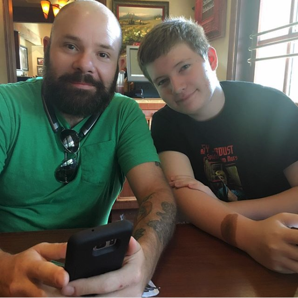 A photograph of two Caucasian males sitting at a table indoors. The man on the left has a shaved head and a full dark beard, wearing a green t-shirt. The younger male on the right has short brown hair and is wearing a black t-shirt.