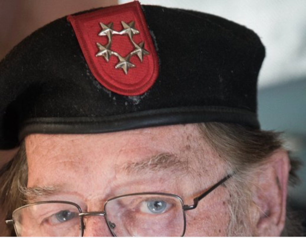 Close-up photograph of a man wearing a black beret with a red patch and five stars, and wire-rimmed glasses.