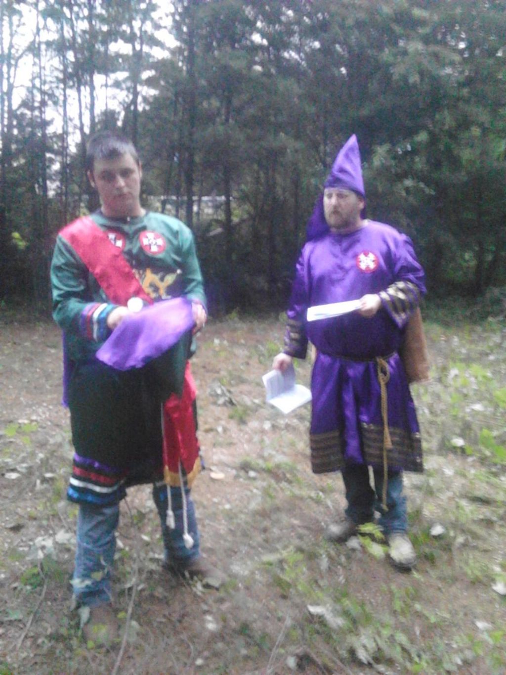 Two men in a wooded area wearing Ku Klux Klan ceremonial robes. One man is in a green robe with a red sash, and the other is in a purple robe with a matching pointed hood.