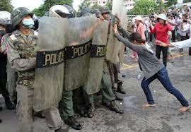 A photograph showing Cambodian riot police with shields standing in a line, while a female protester pushes against them during a demonstration.