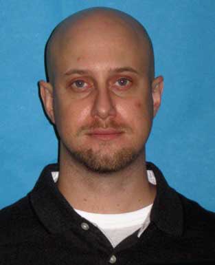 A headshot of a bald Caucasian man with a goatee and light eyes, wearing a dark polo shirt against a plain blue background.