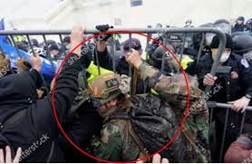A photograph from the January 6th U.S. Capitol attack showing a man in a tactical helmet and camouflage gear at a metal barricade, circled in red. Police officers are visible in the background.