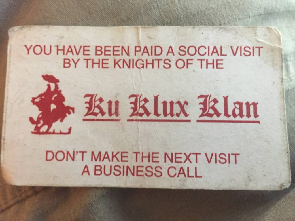 A photograph of a Ku Klux Klan calling card with red text and a graphic of a klansman on a horse. The text reads: 'YOU HAVE BEEN PAID A SOCIAL VISIT BY THE KNIGHTS OF THE Ku Klux Klan DON'T MAKE THE NEXT VISIT A BUSINESS CALL'.