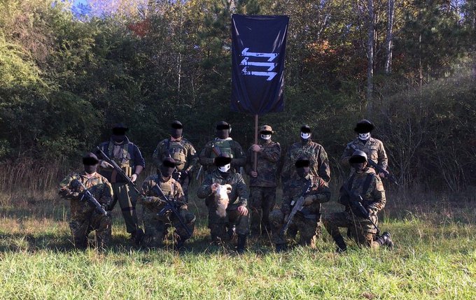 A group of masked individuals in military camouflage and tactical gear posing in a field with firearms and a black flag featuring a white symbol. One individual holds a severed animal head.
