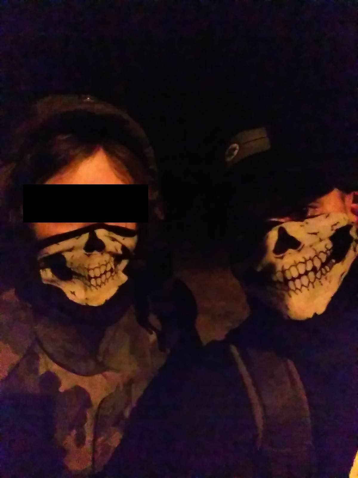 A low-light photograph of two individuals wearing skull-patterned face masks. One person's eyes are obscured by a black censor bar. They appear to be wearing dark clothing and headgear.