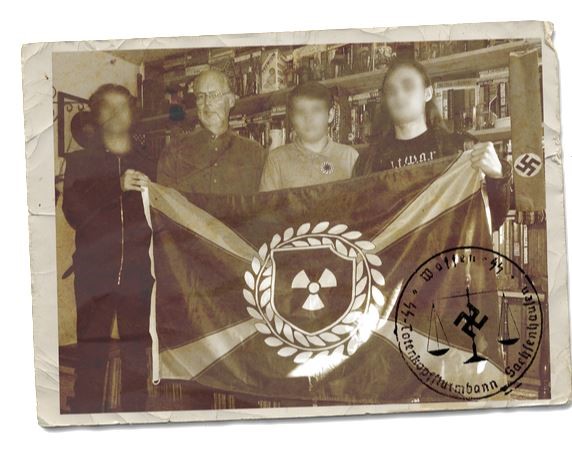 A sepia-toned, aged photograph showing four individuals holding a flag with a radiation symbol (Atomwaffen Division). The second person from the left is a bald Caucasian male with glasses. The faces of the other three individuals are blurred. Nazi symbols, including a swastika banner and a circular stamp with German text, are visible.