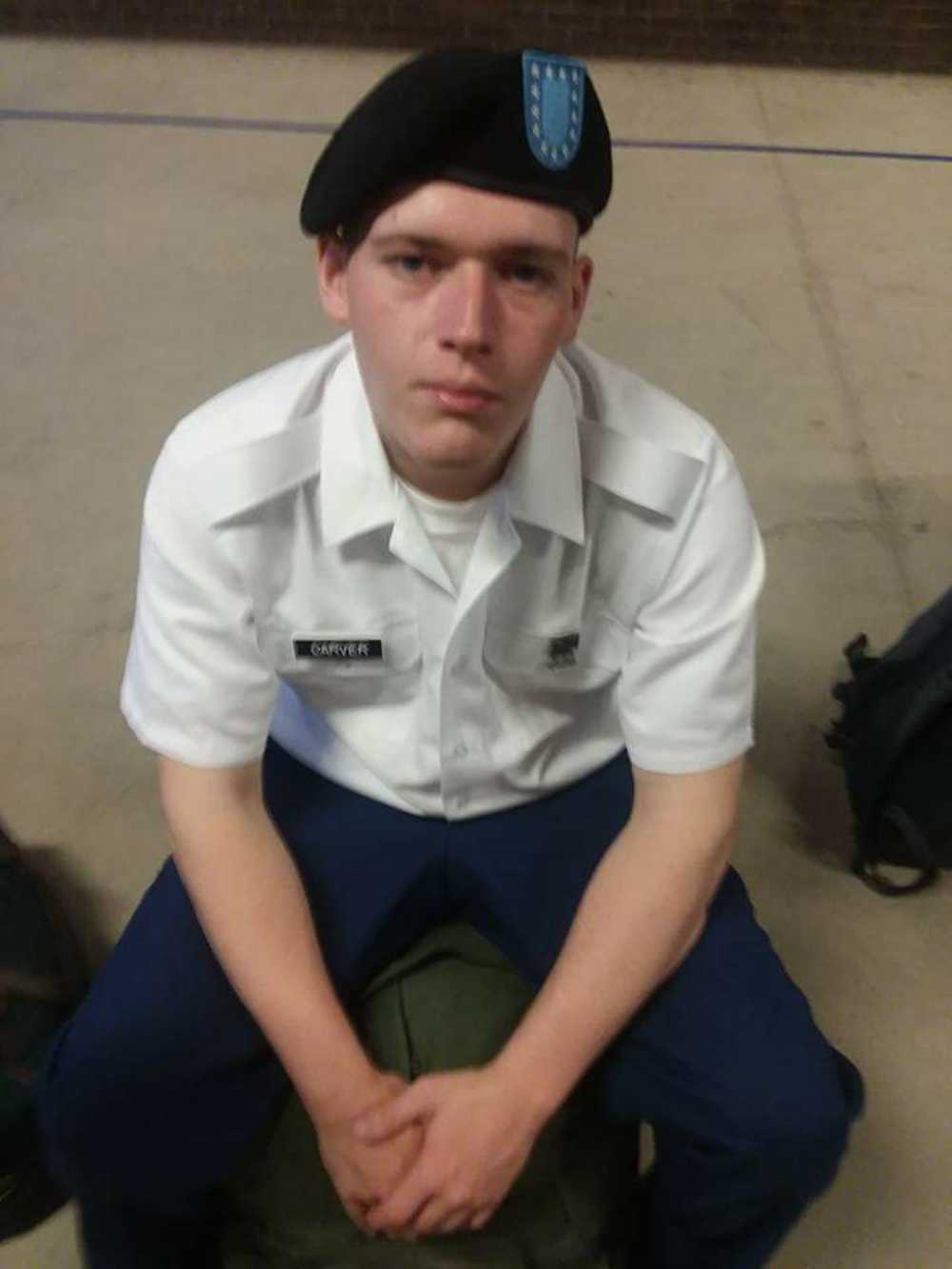 A photograph of a young man in a U.S. Army uniform, wearing a white short-sleeved shirt with a name tag that reads "CARVER" and a black beret. He is sitting down with his hands clasped.