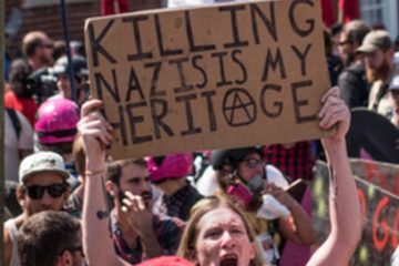 A protester in a crowded street holds a cardboard sign that reads 'KILLING NAZIS IS MY HERITAGE' with an anarchy symbol drawn on it. Other protesters with helmets and sunglasses are visible in the background.