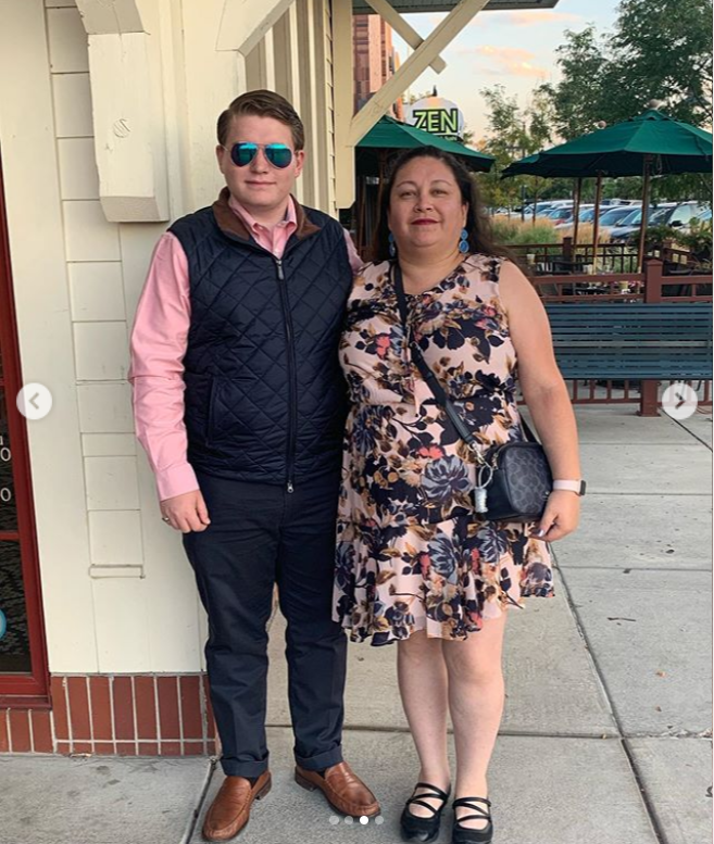 A man with brown hair wearing a pink shirt, navy quilted vest, and sunglasses stands next to a woman in a floral dress on a sidewalk.