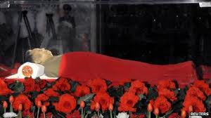 A photograph of a deceased North Korean leader lying in state inside a glass casket, covered with a red blanket and surrounded by red flowers.