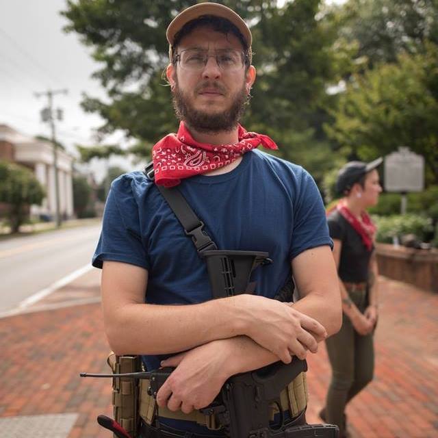 A man with a beard, glasses, and a tan cap stands in a street holding a rifle. He wears a red bandana and a blue t-shirt. Another person is visible in the background.