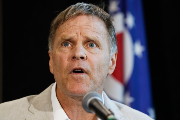 A close-up photograph of a middle-aged Caucasian man with short, light brown hair and blue eyes, speaking into a microphone. He has a concerned or intense expression. A flag with stars and a red circle is visible in the background.
