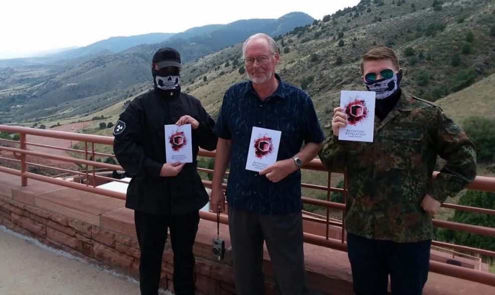 Three men stand at an outdoor overlook with mountains in the background. Two men wear skull masks and tactical gear, holding pamphlets titled 'REVISITING REVELATION'. The man in the center is unmasked, wearing a blue patterned shirt and glasses, also holding a pamphlet.