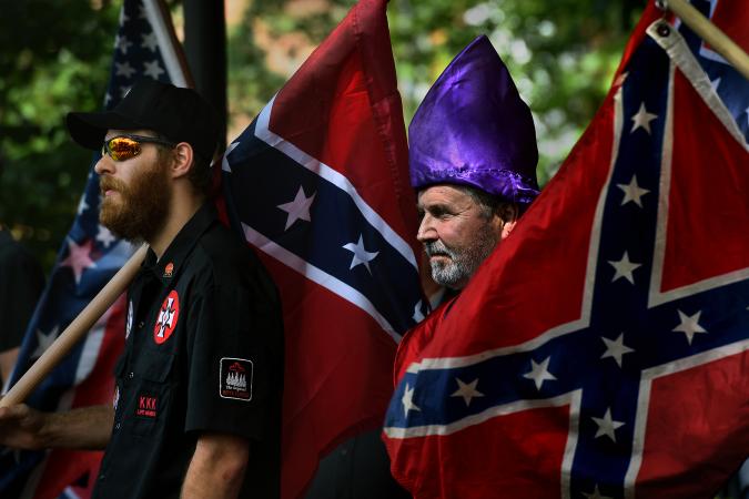 Two men at a white supremacist rally holding Confederate flags. One man wears a black shirt with KKK patches and a baseball cap, while the other wears a purple KKK hood.