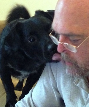 A close-up photograph of a bald Caucasian man with a grey beard and glasses lying down while a black dog, identified as Lamont in the context, nuzzles his face.