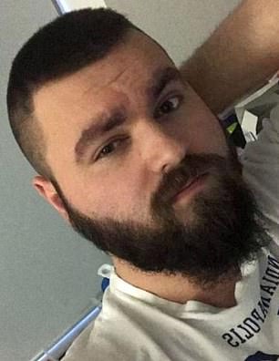 A close-up photograph of a Caucasian man with a short buzz cut and a full dark beard, wearing a white t-shirt.