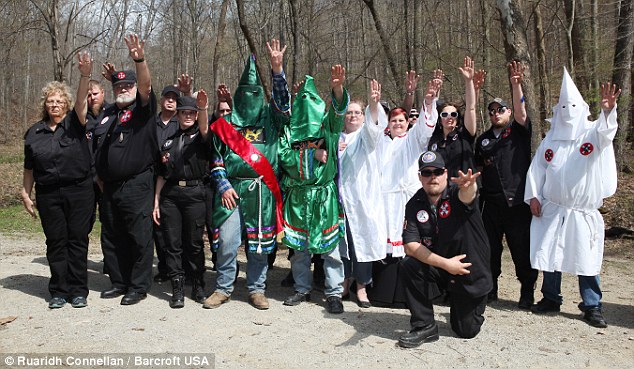 A group of individuals associated with the Ku Klux Klan posing in a wooded area. Some wear traditional green and white robes with hoods, while others wear black shirts with patches. Many are performing a one-armed salute.