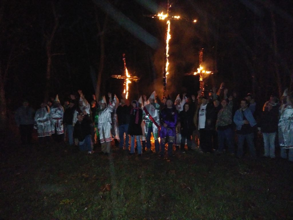 A group of people, some wearing KKK robes and hoods, standing in front of three burning crosses at night. Many individuals have their arms raised in a salute.