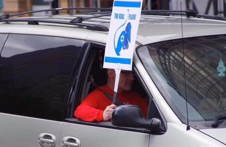 A man in a red shirt and glasses sits in a silver minivan, holding a sign out the window with antisemitic imagery and the text 'THE REAL PLAGUE' during a protest.