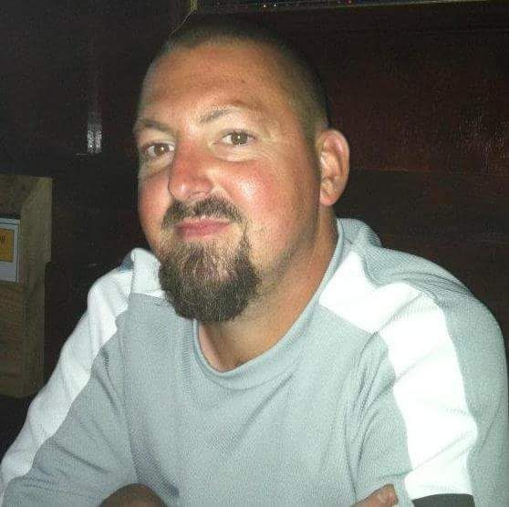 A close-up photograph of a Caucasian man with a buzz cut and a goatee, wearing a grey and white athletic shirt.