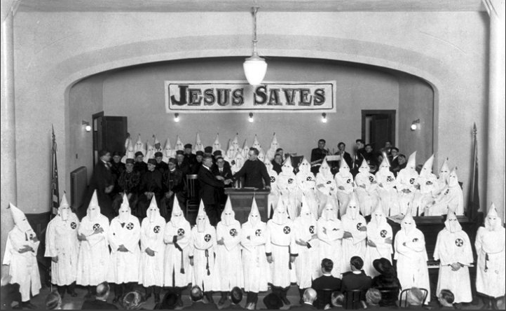A historical black and white photograph of a large group of Ku Klux Klan members in white robes and pointed hoods standing on a stage. A banner above them reads 'JESUS SAVES'. Several men in dark suits are also visible on the stage.