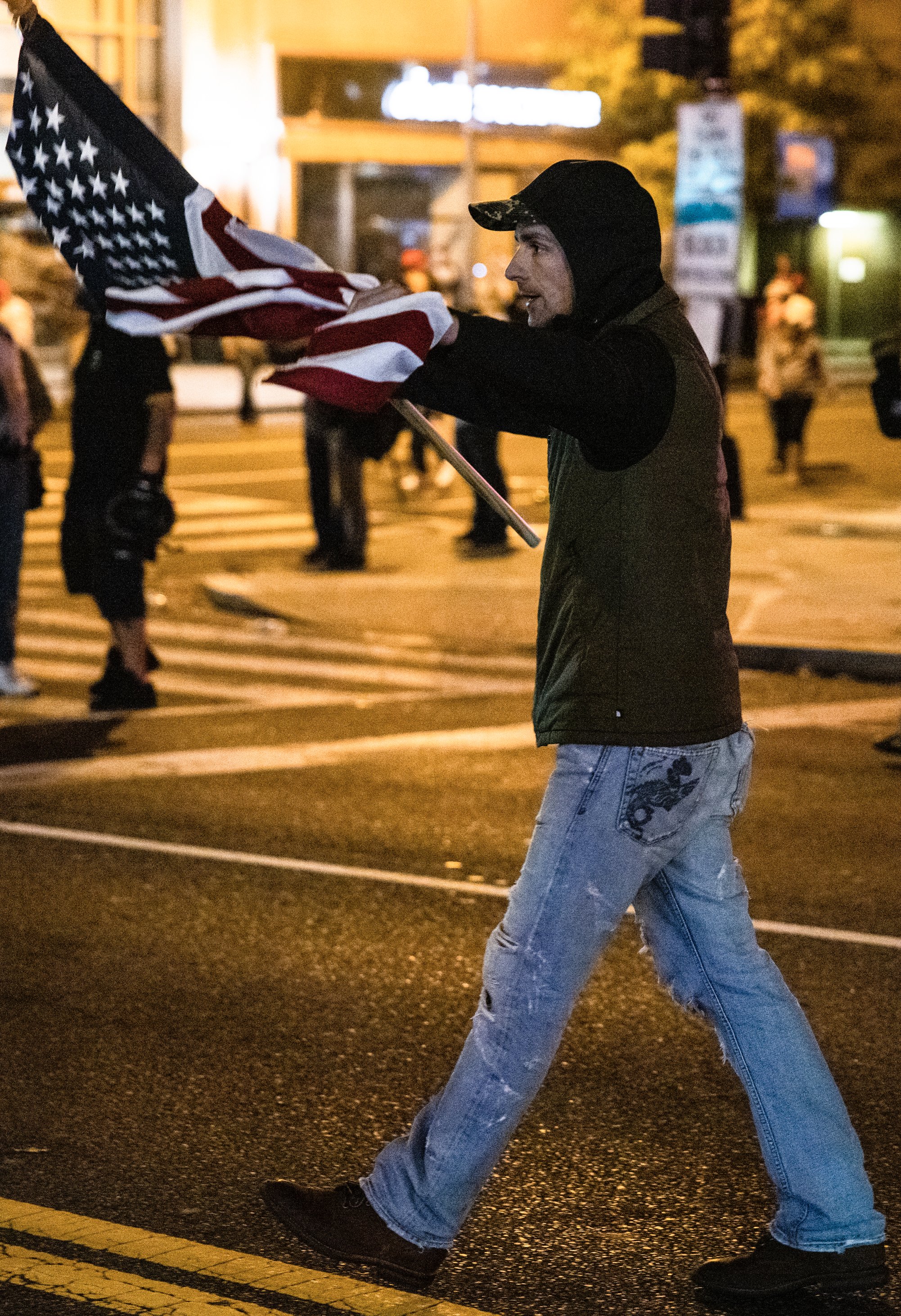 A man in a green vest and baseball cap walks across a city street at night carrying an American flag.