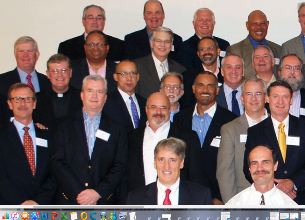 A screenshot of a group photograph featuring approximately twenty men in formal business attire, including one man wearing a Catholic clerical collar.