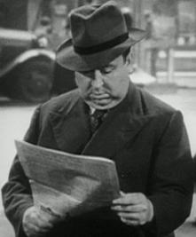 A black and white photograph of a heavy-set man in a suit and fedora reading a newspaper on a street.