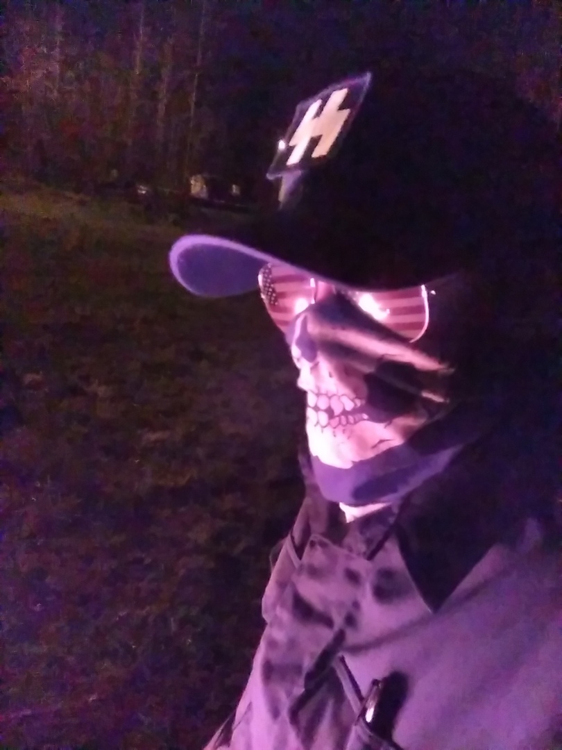 A person wearing a black baseball cap with a white symbol, American flag aviator sunglasses, and a skull-patterned face mask, illuminated by a purple light source at night.