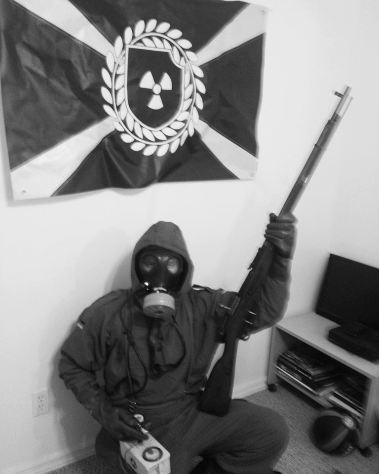 A black and white photograph of a person wearing a gas mask and a hooded suit, holding a rifle and a Geiger counter. Behind them hangs a flag associated with the Atomwaffen Division, featuring a radiation symbol.