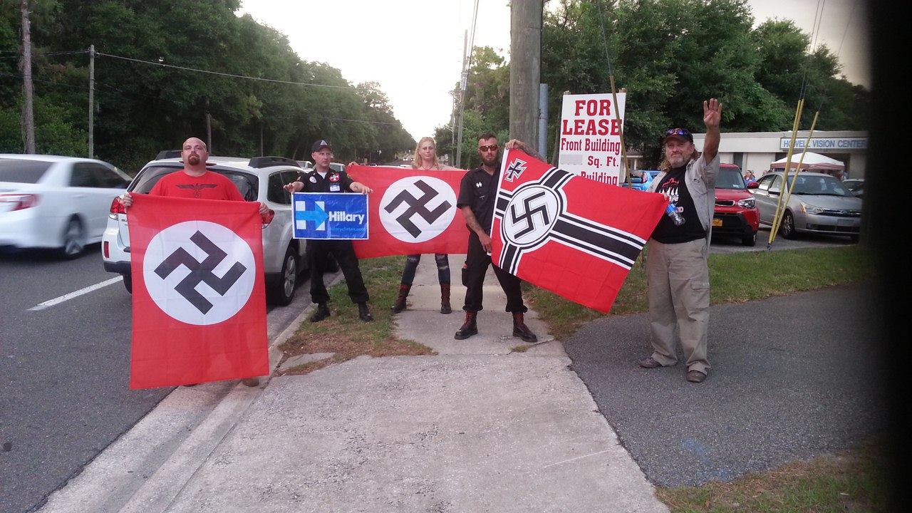 A group of five individuals standing on a sidewalk next to a road, holding three large red flags with black swastikas in white circles and a blue Hillary Clinton campaign sign. One man on the far right is performing a Nazi salute.