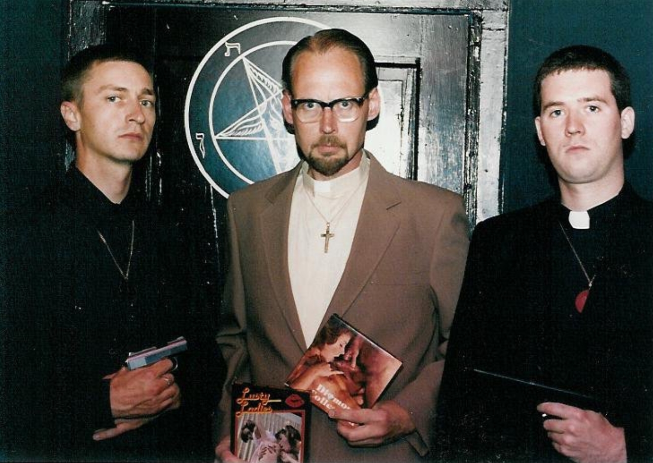 Three men posing in front of a door with a pentagram symbol. The man in the center wears a tan suit and clerical collar, holding adult magazines. The two men flanking him wear black and hold handguns.