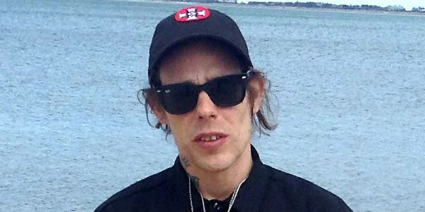 A photograph of a man wearing a black baseball cap with a KKK-style logo, sunglasses, and a black shirt, standing in front of a body of water.