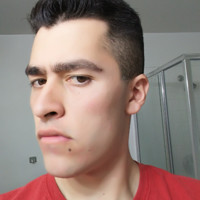 A close-up photograph of a young man with dark hair and a mole on his chin, wearing a red shirt.