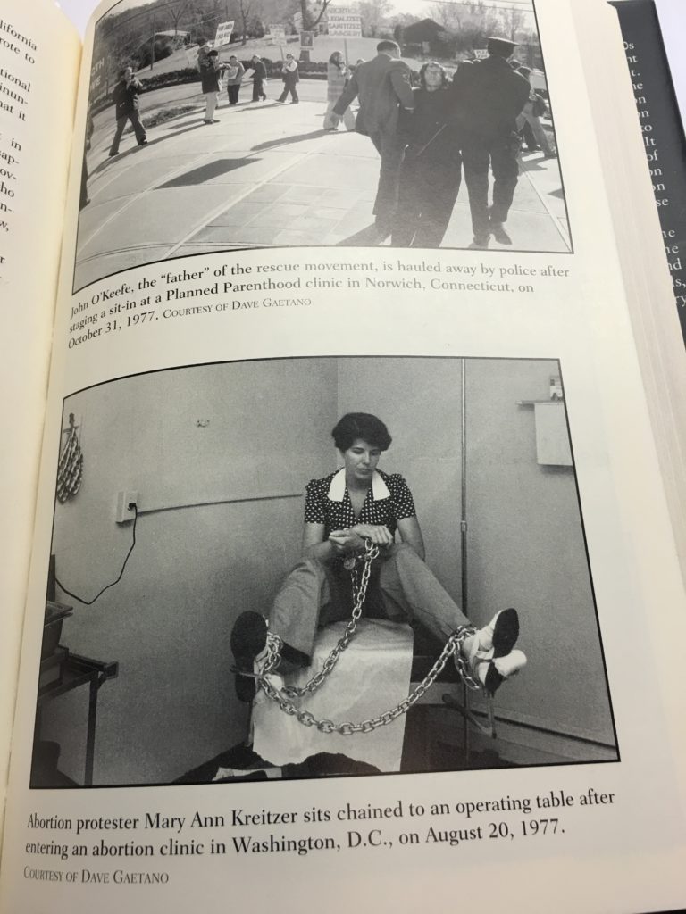 A photograph of a book page featuring two black and white images of anti-abortion protesters from 1977. The top image shows John O'Keefe being arrested by police. The bottom image shows Mary Ann Kreitzer chained to an operating table.