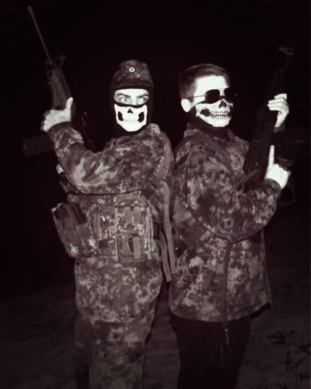 A black and white photograph of two individuals in camouflage military-style clothing, wearing skull-patterned face masks and holding assault rifles. One individual on the right has visible short hair and is wearing sunglasses.