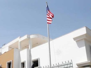 A photograph of an American flag flying on a pole in front of a white, multi-story building, likely a U.S. Embassy, against a clear blue sky.
