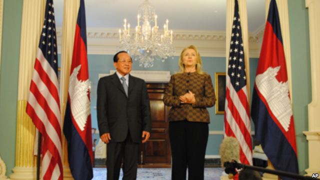 Hillary Clinton and a Cambodian official standing between the United States and Cambodian flags in a formal room with a chandelier.