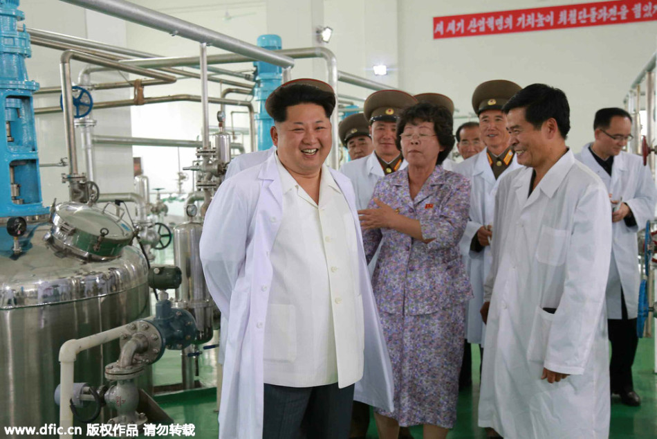 Kim Jong Un, wearing a white lab coat over a light-colored shirt, smiles broadly while standing in what appears to be a factory or laboratory. He is surrounded by other individuals, some in lab coats and others in military-style uniforms or civilian clothes, all of Asian ethnicity. Large industrial machinery and pipes are visible in the background. A red banner with Korean text hangs on the wall.