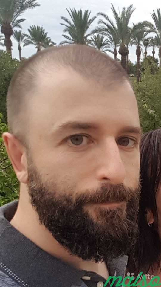 A close-up photograph of a Caucasian man with a buzz cut and a full dark beard, standing outdoors with palm trees in the background.